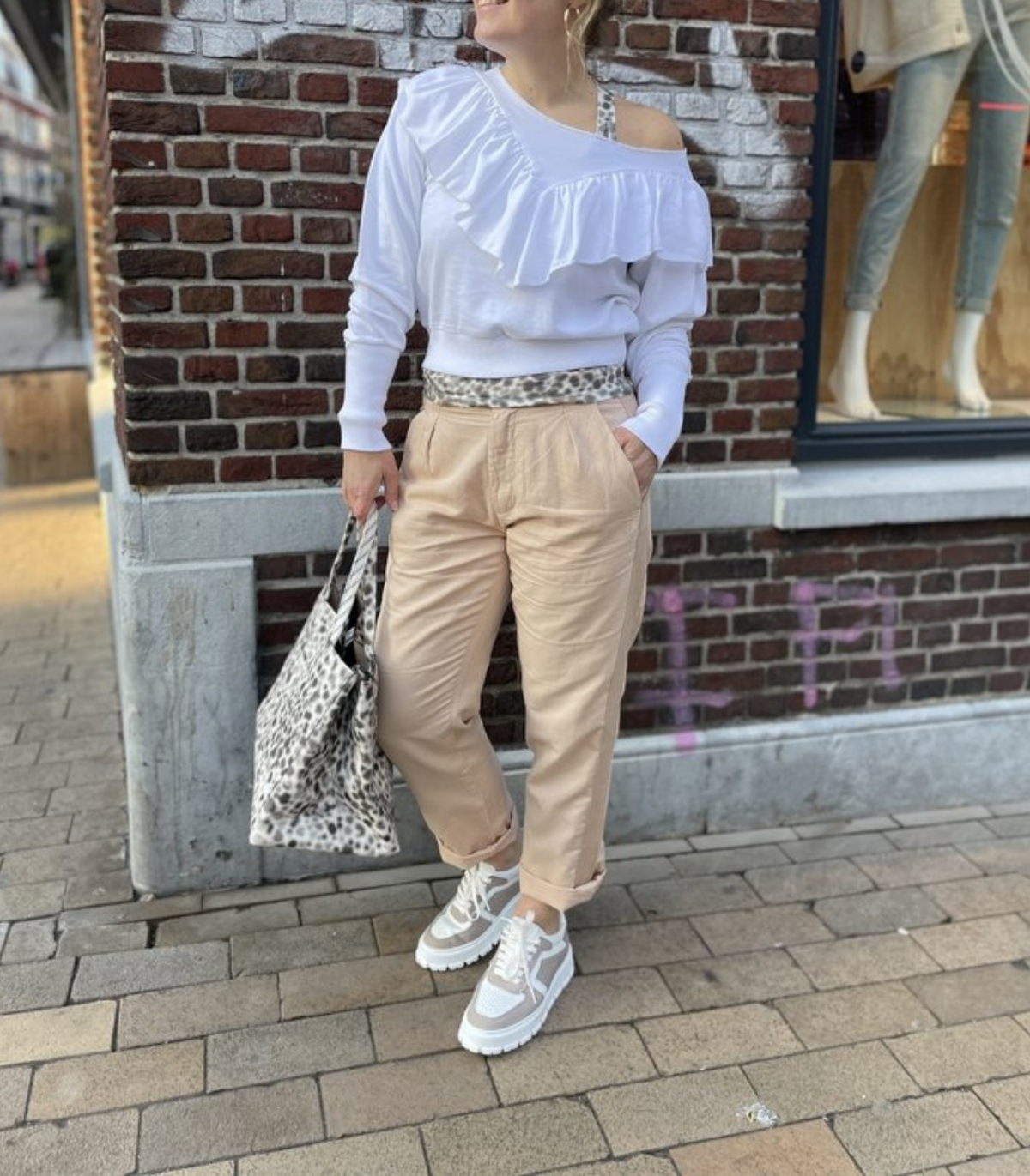 Frau in weißem Rüschenpullover, beigen Hosen, Leopardenmuster-Tasche und Sneakers auf Straße.