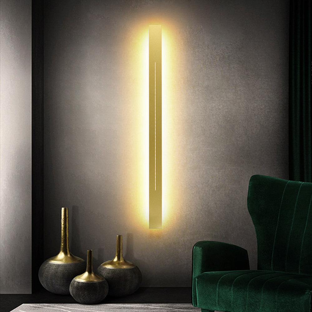 Moderne LED-Wandleuchte, vertikal, goldfarben, beleuchtet, neben grünem Samtsessel.