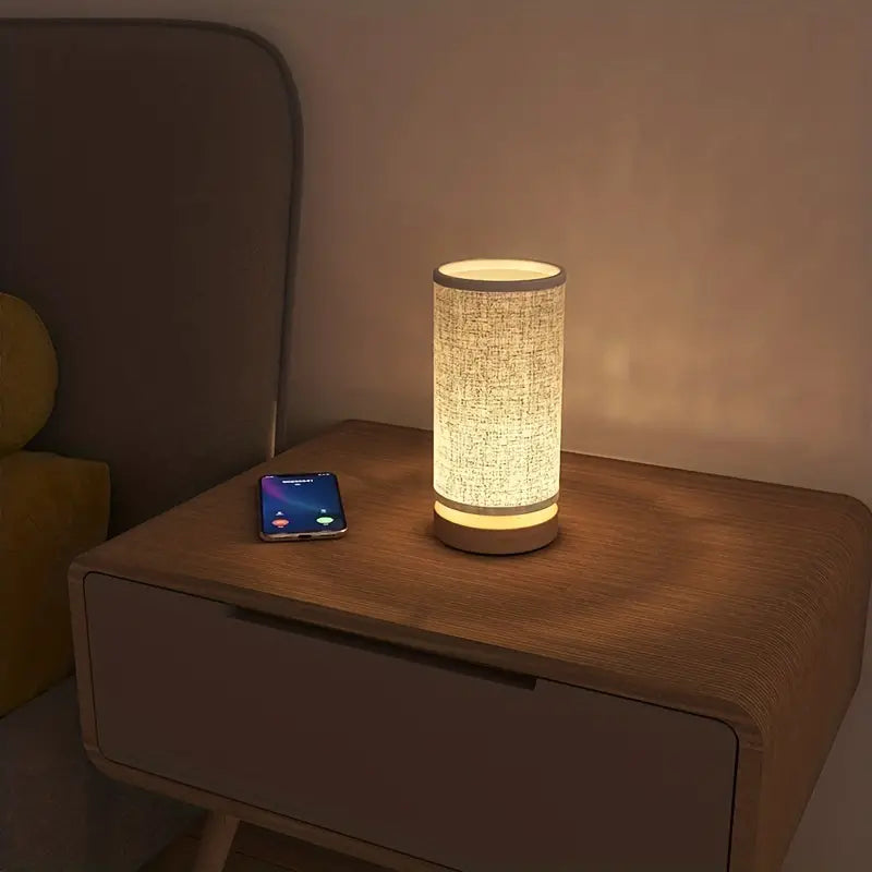 Nachttischlampe aus Stoff, zylindrisch, warmes Licht, auf Holztisch neben Smartphone.