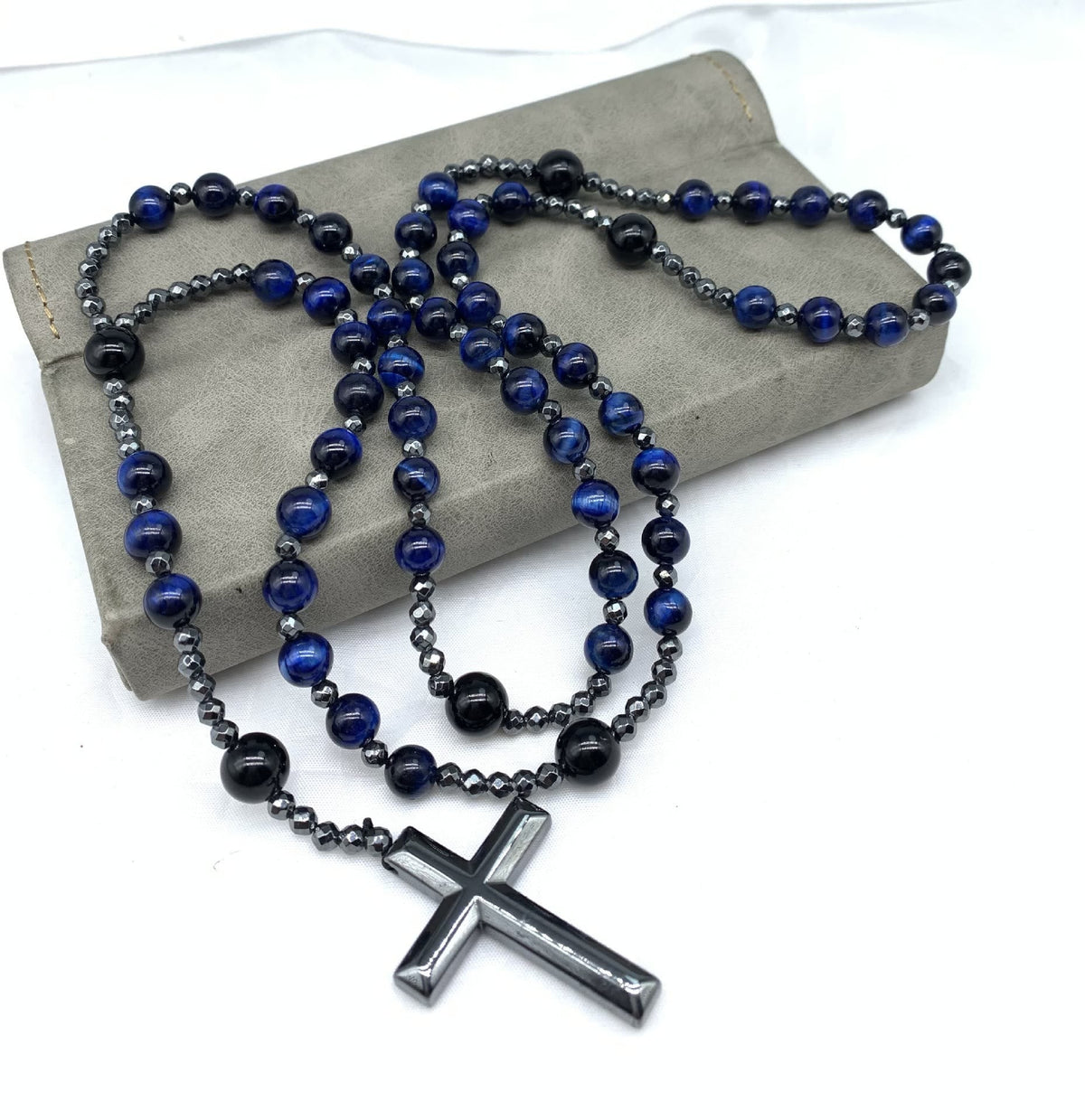 Blau-schwarze Perlenkette mit silbernem Kreuzanhänger auf grauem Hintergrund, modisches Accessoire.