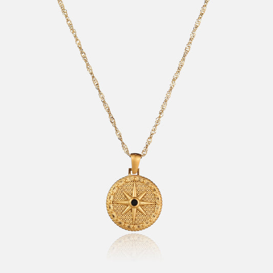 Guldfärgat halsband med rund hängsmycke format som en åttauddig stjärna med en liten svart sten i mitten, sett framifrån mot ljus bakgrund. 1