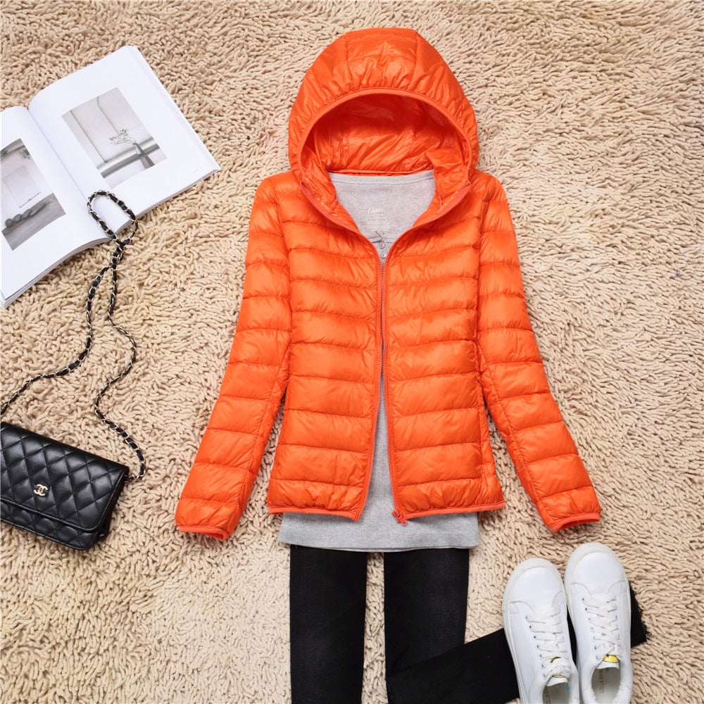 Orangefarbene Daunenjacke mit Kapuze auf Teppich, neben schwarzer Tasche und weißen Sneakers.