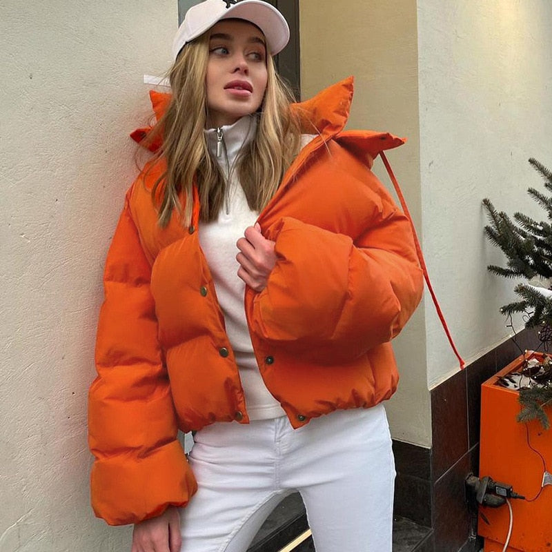 Frau in orangefarbener Daunenjacke, weißem Pullover und weißer Hose, modisch, urban.