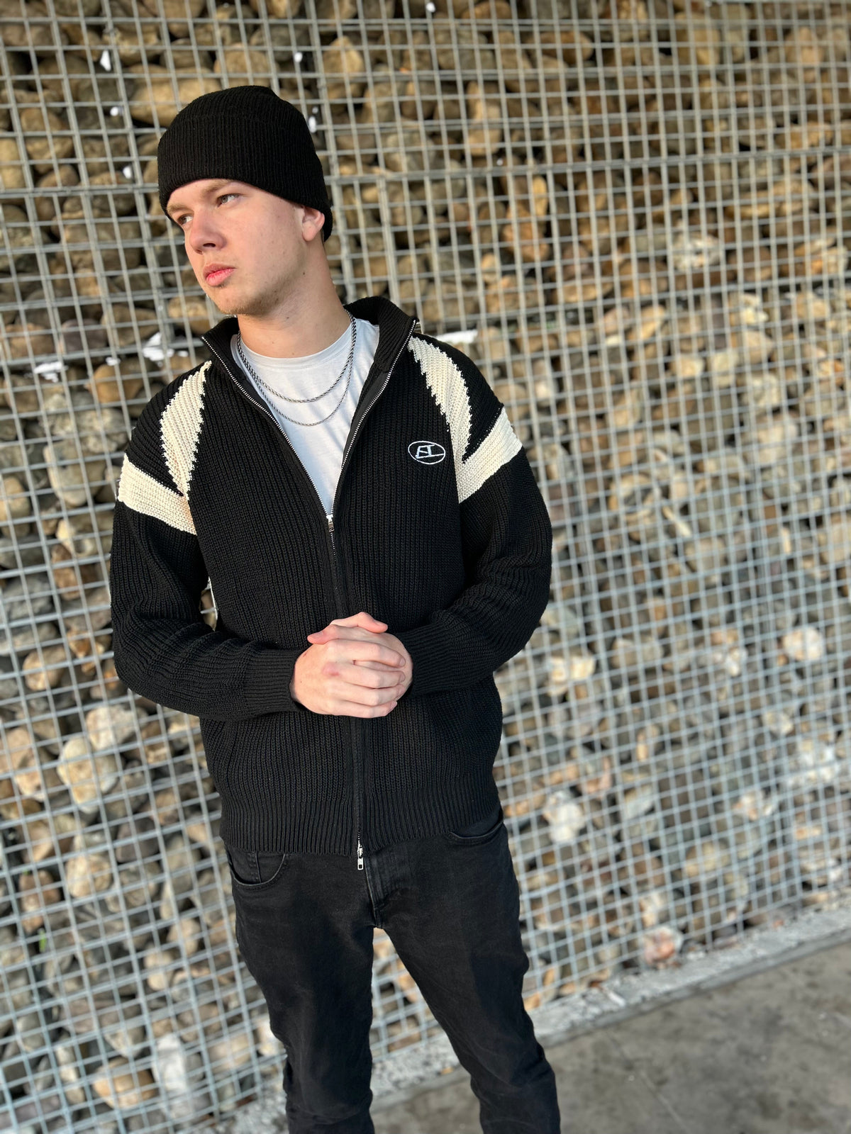 Mann in schwarzer Strickjacke mit weißen Streifen und Mütze vor Steinwand.