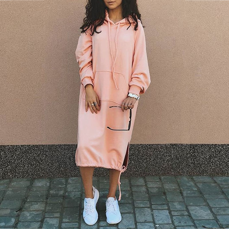 Frau in rosa Hoodie-Kleid, weiße Sneaker, hält Sonnenbrille, lässiger Streetstyle-Look.