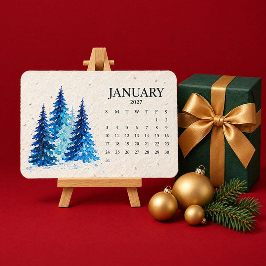 Januarikort med blå granar på ett träbord-staffli, bredvid en grön julklapp med guldfärgad rosett och julkulor 2