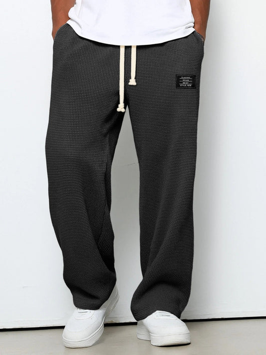 Man bär mörkgrå våffelstickade byxor med vida ben och krämvit dragsko, framifrån med händerna i fickorna och vita sneakers. 1