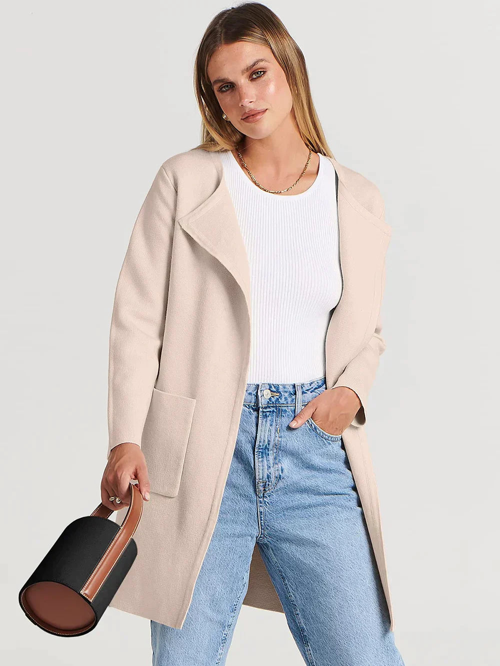 Frau in beigem Wollmantel, weißem Pullover, blauer Jeans, hält schwarze Handtasche.