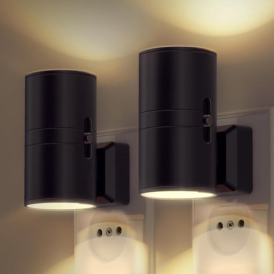 Två svarta cylinderformade nattlampor insatta i vägguttag, med varmt ljus som sprids uppåt och nedåt längs väggen. 1