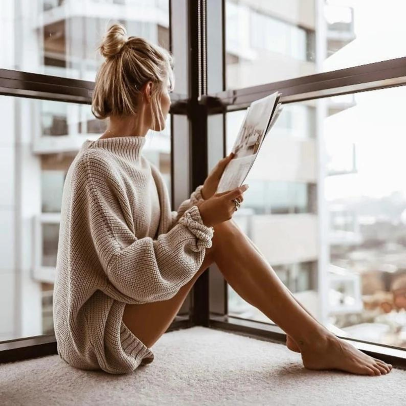 Frau in beigem Strickpullover liest Zeitschrift am Fenster, moderne Wohnung, gemütlich.