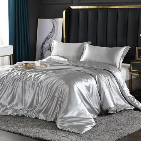 Luxuriöses silbernes Satin-Bettwäsche-Set auf modernem Bett mit Kopfteil und grauem Teppich.
