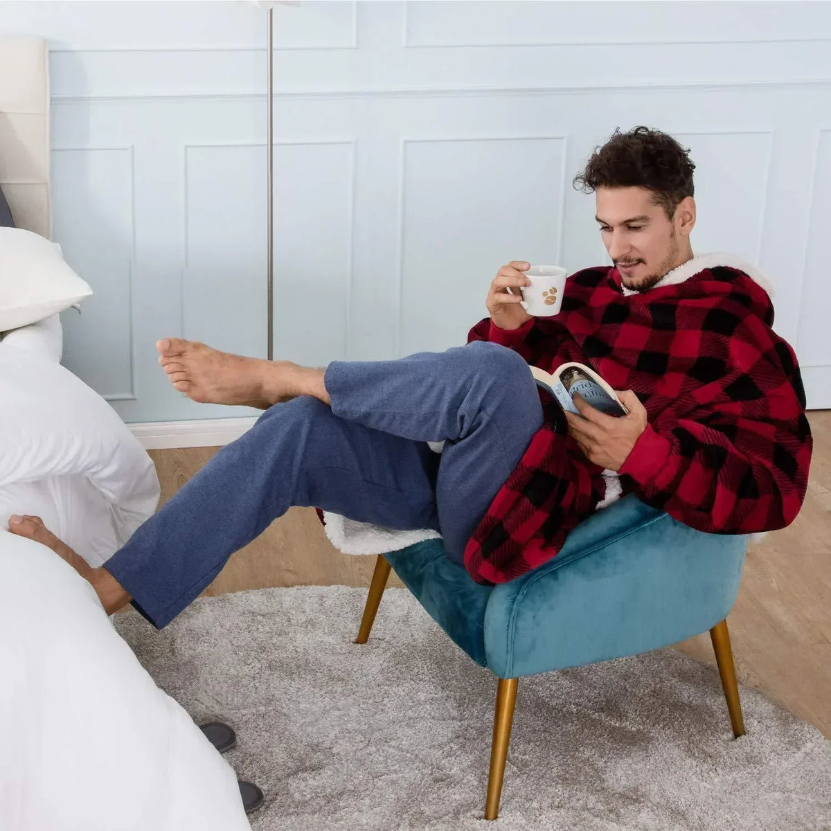 Mann in rotem Karo-Sweatshirt liest Buch, trinkt Kaffee, sitzt auf blauem Sessel im Schlafzimmer.
